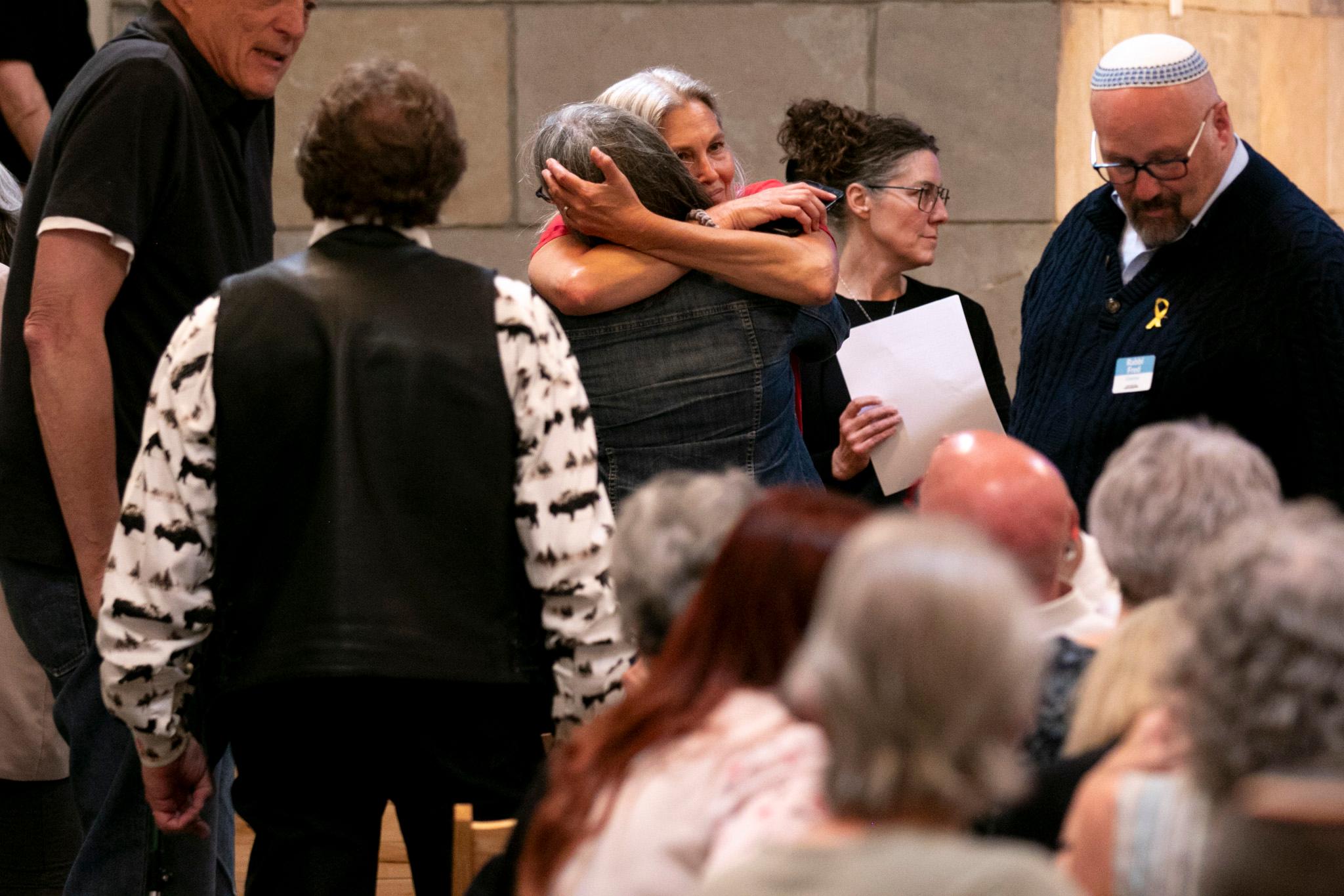 A group of people are gathered in a room, with one woman hugging another woman. The woman hugging the woman is wearing a pink shirt. The room has a beige wall and a wooden floor.