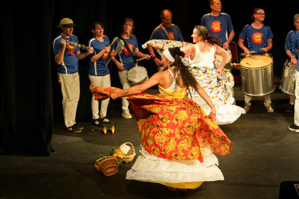 Two dancers spin and hold out their long, colorful skirts. A band performs live behind them.