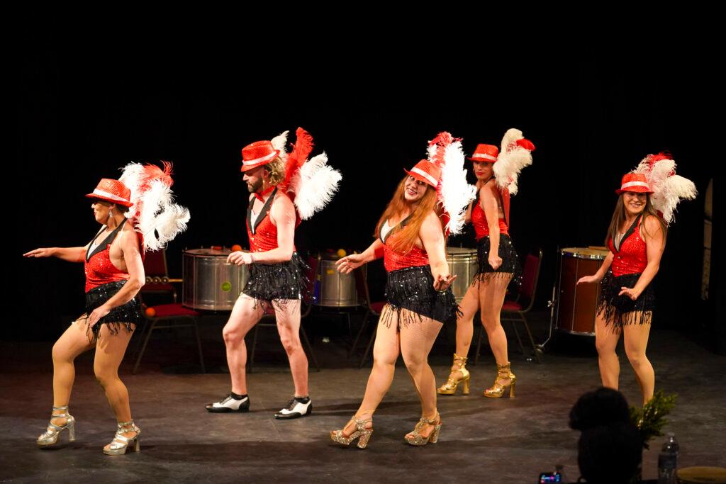 Five dancers in black shorts with fringe, red halter tops and red hats, plus large red and white feathers off their backs, perform for the competition’s judges.