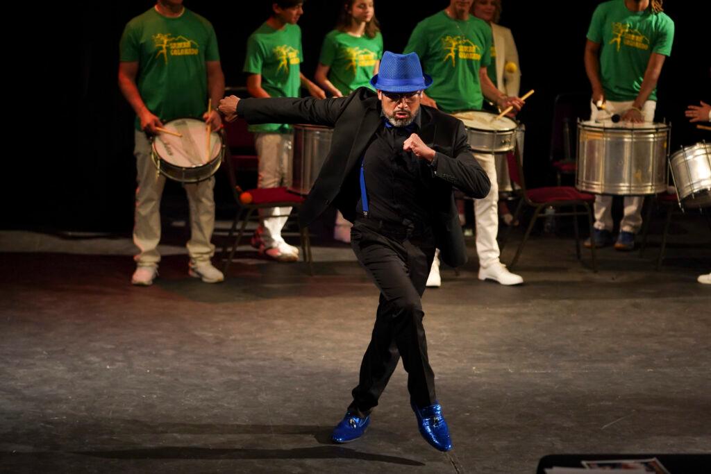 A dancer in a black suit with a royal blue hat and royal blue shimmery shoes dances the samba. A band plays live behind him.
