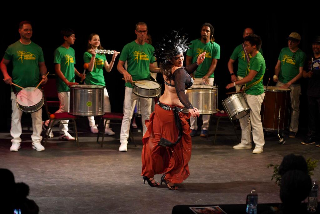 A dancer wearing flowy, red pants, a black bra top and a black headdress twists her hips toward the audience. A live band plays behind her.