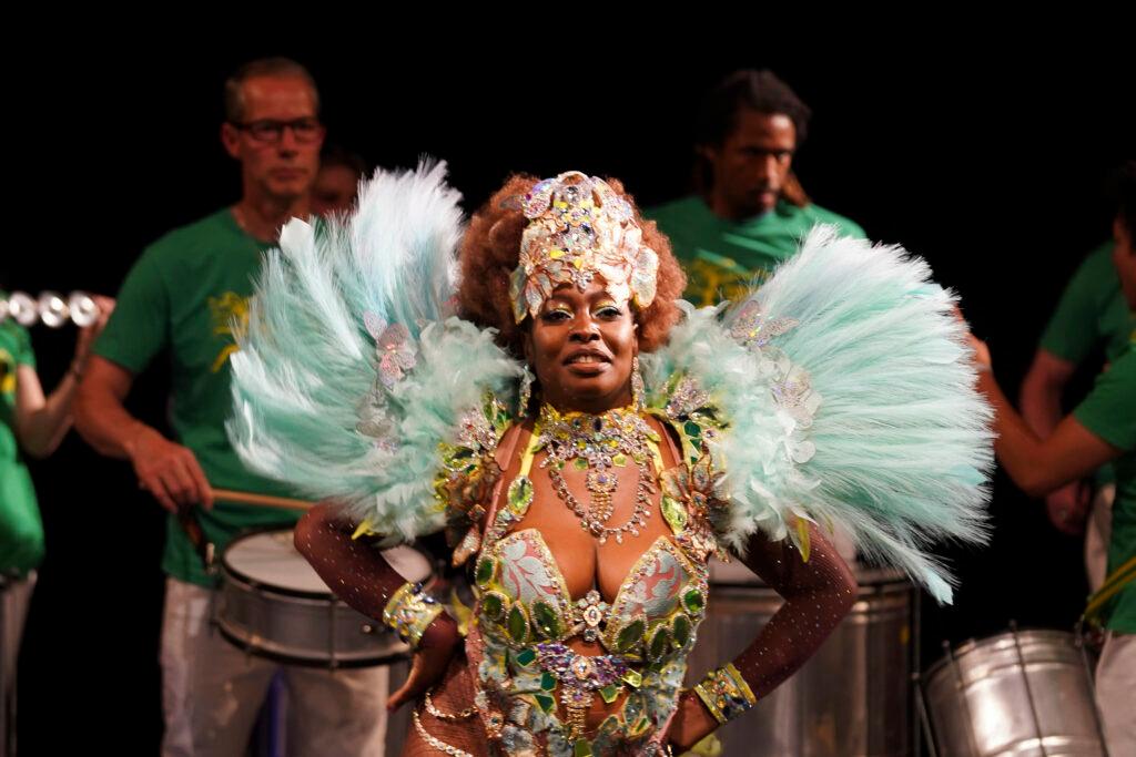 A samba dancer with big teal feathers off her shoulders and an elaborate headdress shimmies onstage. Several drummers are behind her.