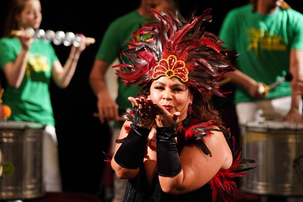 A dancer in a black fringe dress holds her hands up to her face as she blows a kiss. She wears black arm bands and a red and black feathered headdress. A band plays live behind her, many of the musicians are playing drums.