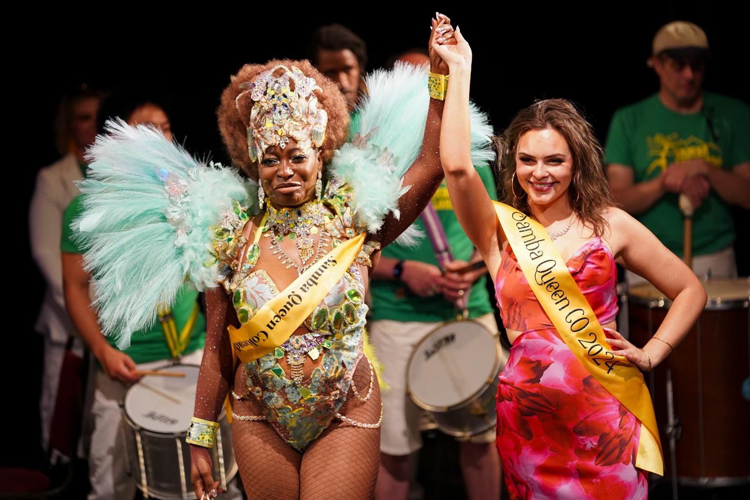 A dancer with a yellow sash that says “Samba Queen Colorado 2025” and big teal feathers coming up her shoulders holds hands with another dancer, who wears a sash that says “Samba Queen Colorado 2024.” Both are smiling triumphantly.