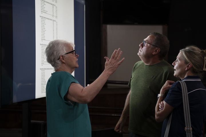 An older woman speaks to a man and an woman who are looking over her shoulder at a large screen displaying election results