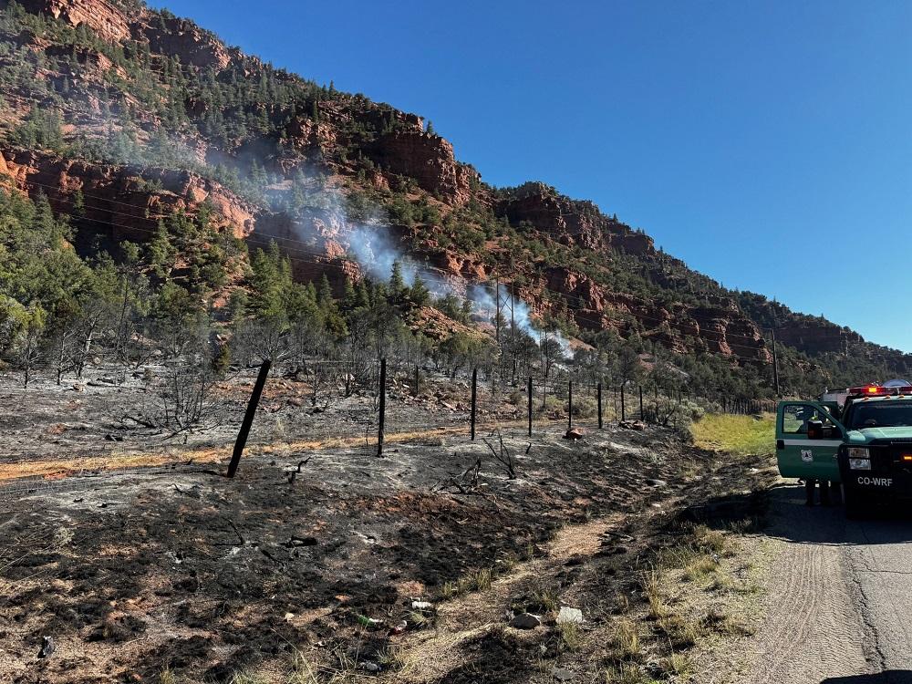 A burned landscape is seen along the edge of a road with smoke rising from the ground in one spot in the background.
