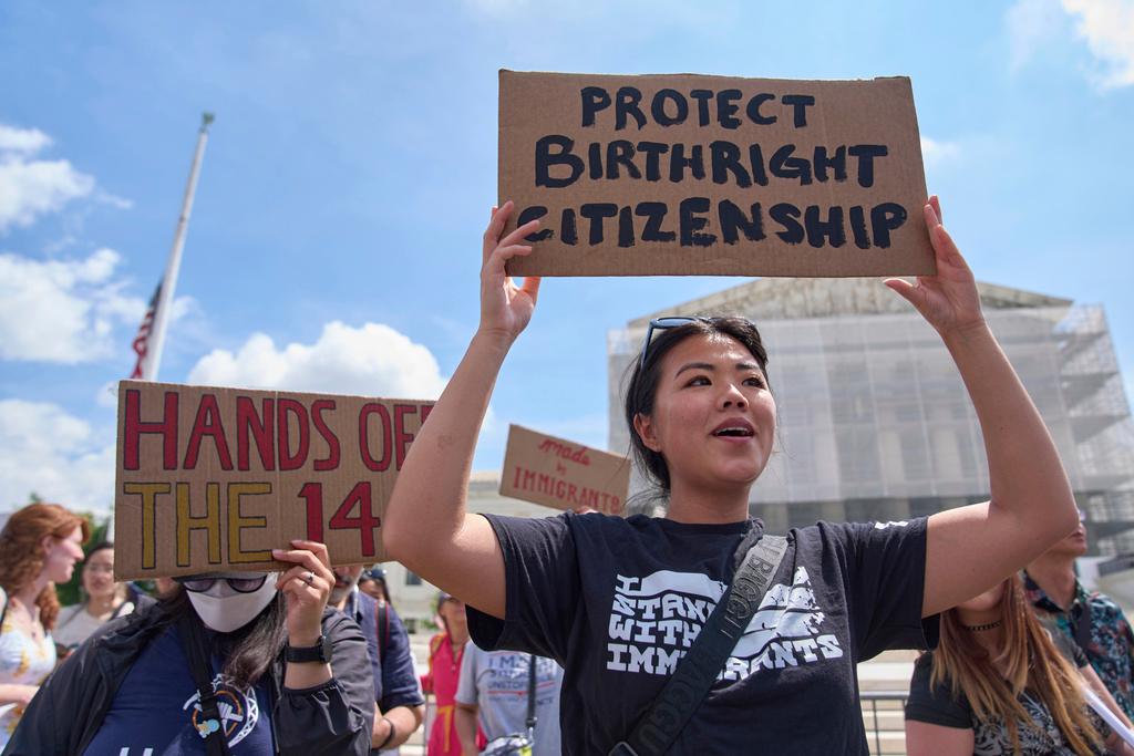 A Chinese woman wearing a black t-shirt with a slogan supporting immigrant rights holds a cardboard sign reading "PROTECT BIRTHRIGHT CITIZENSHIP." Behind her are other demonstrators holding their own protest signs.