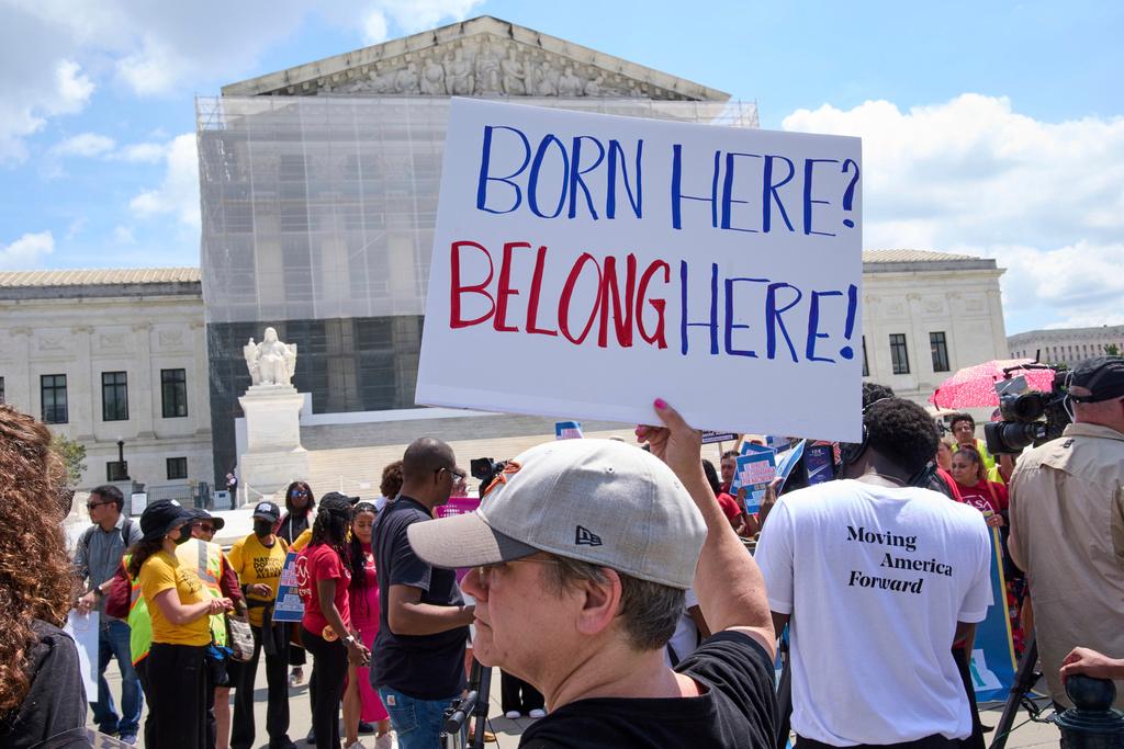 A white woman with short hair and a baseball cap holds a sign that reads "BORN HERE? BELONG HERE!" in a crowd of other demonstrations in front of the Supreme Court building.