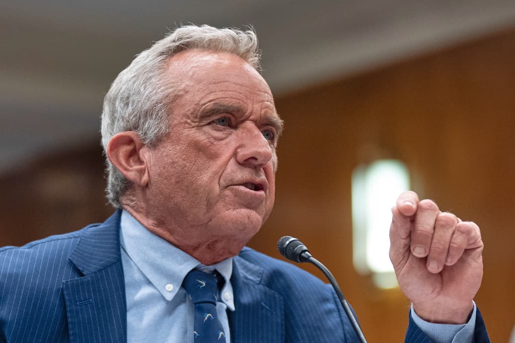 A man in a suit is speaking into a microphone. He is wearing a blue tie and has gray hair.