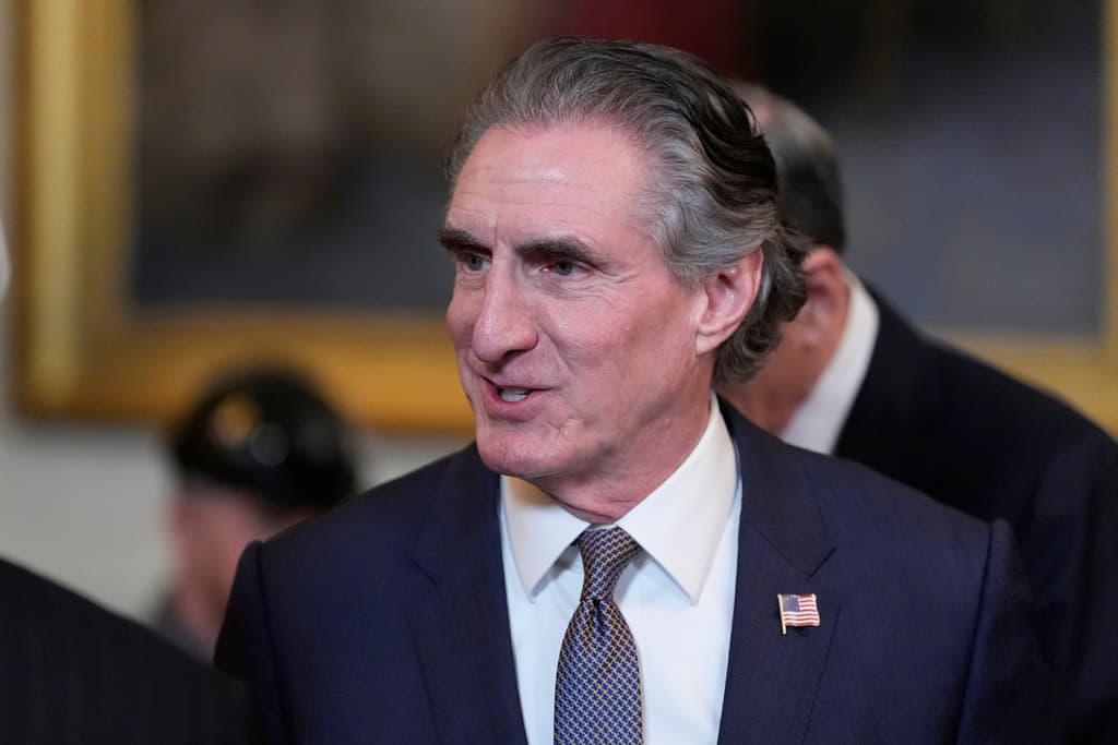 A man in a dark blue suit and a white shirt is the main subject of the image. He is wearing a blue tie and a pin on his suit.