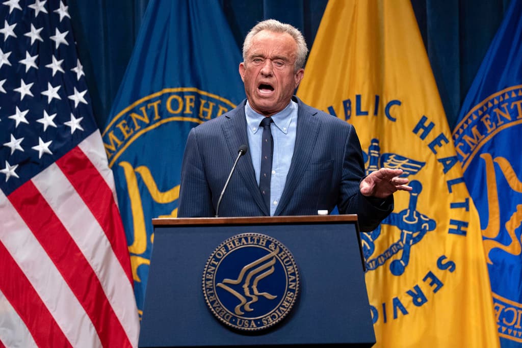 A man in a dark suit and tie stands behind a podium with a microphone, giving a speech. The podium is has multiple flags and the words &quot;United States Army&quot; are displayed.