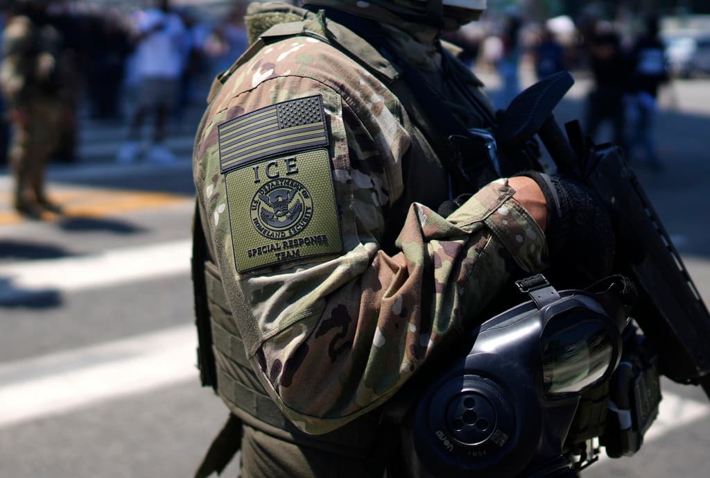 A person in a military uniform stands on a street, holding a gun. The uniform is camouflaged, and the person's face is obscured. The background is blurred.