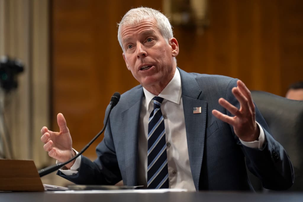 A man in a gray suit and blue tie is seated at a desk, gesturing with his hands. He is speaking into a microphone placed on the desk. The background features a wooden panel and a camera