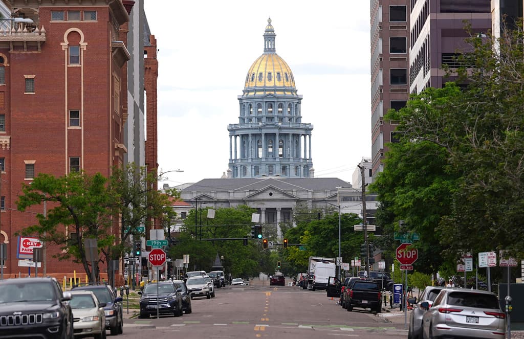 The image depicts a city street with a large, tall building featuring a gold dome. The street is lined with various cars parked on both sides, and there are multiple traffic lights at different points along the road.