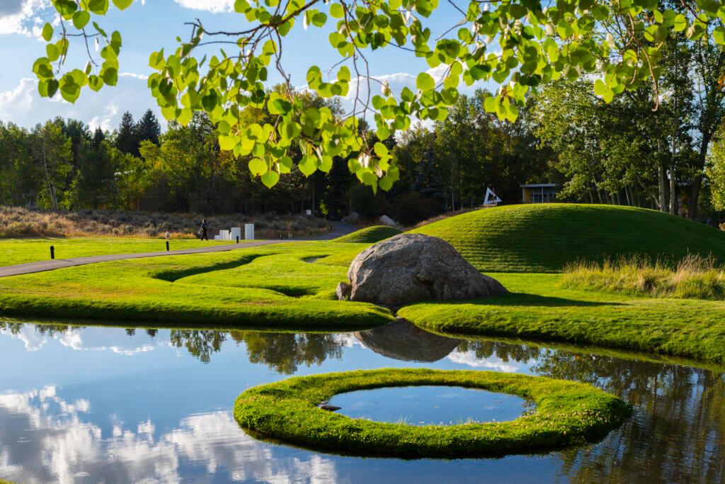 A sculpted landscape featuring a grass mound with a moat around it, a large rock, and a pnd with a grass circle inside it, encasing another, tiny pond.