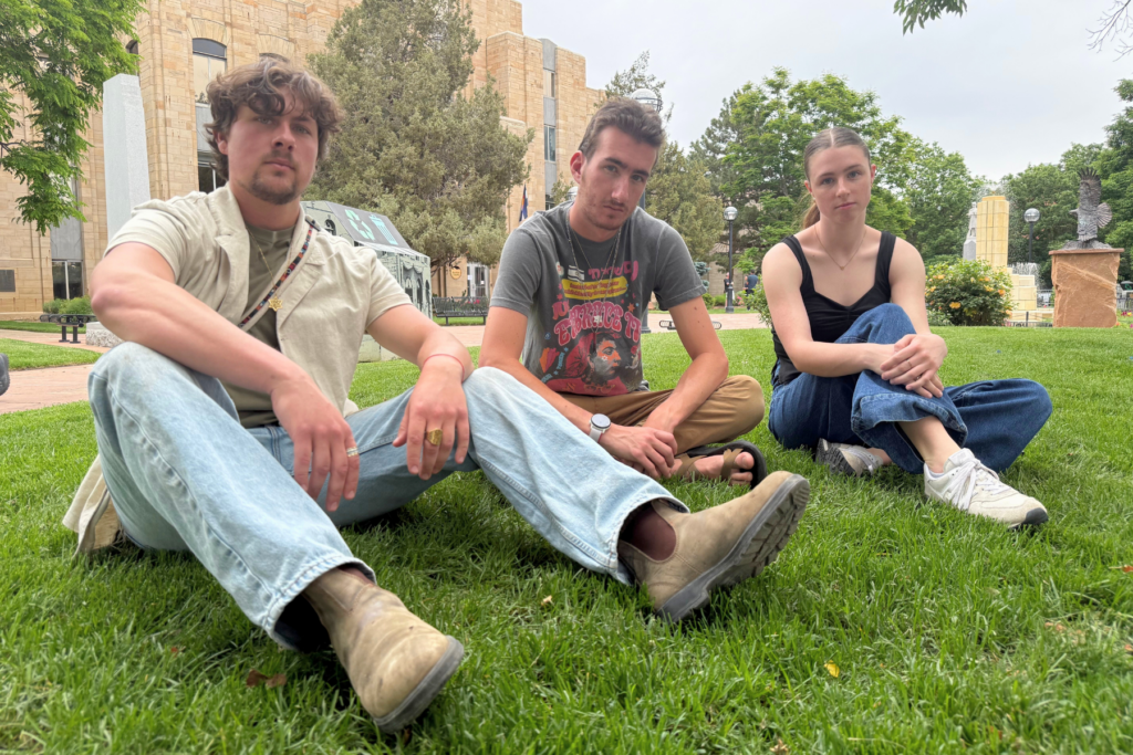 Three young adults sit on green grass with the Boulder County courthouse and trees in the background. Two men sit cross-legged; the woman sits with her knees up. All have neutral expressions and wear casual clothes.
