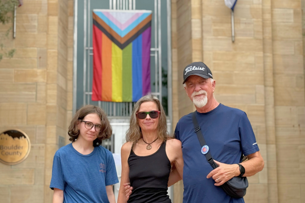 Three people stand smiling in front of a building with a large Progress Pride flag hanging above the entrance. The building is marked Boulder County on a plaque to the left.