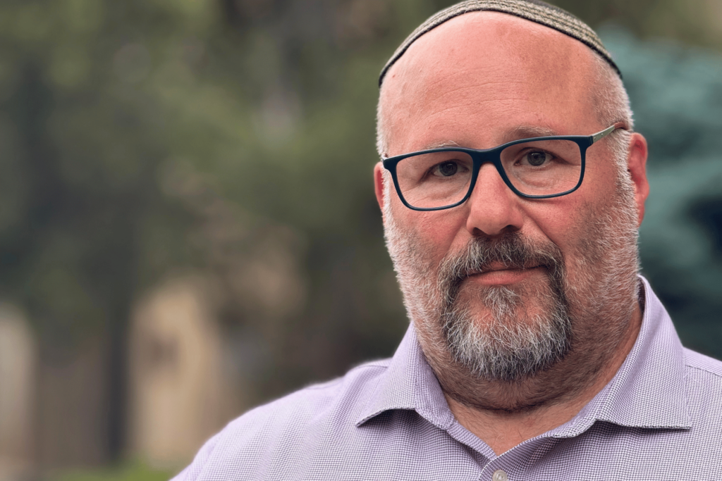 A middle-aged man with a trimmed gray beard, glasses, and a kippah, wearing a light purple shirt, stands outdoors with a blurred green background.