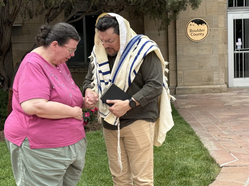 A man and a woman hold hands praying near a patch of grass. The man is wearing a traditional head covering and holding a bible.