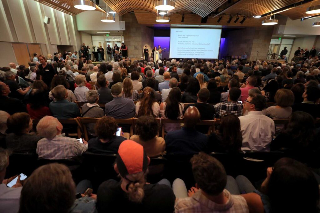A large group of people are gathered in a room, watching a presentation on a screen. The audience is seated on chairs, with some standing. The room has a mix of people, with some individuals closer to the front and others further back.