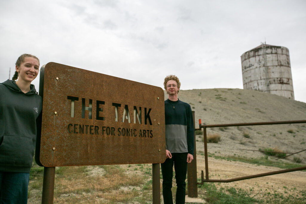 Holly McMahon, right, stands next to a sign that reads "THE TANK CENTER FOR SONIC ARTS" with Asher Dobrin left of the sign and the tank visible on a hill behind them.