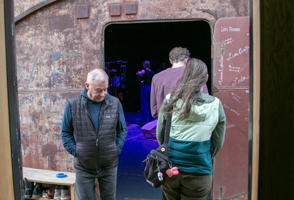A man stands outside the door toa large, metal tank as two people enter the tank. Someone inside playing a windchime is partially visible.