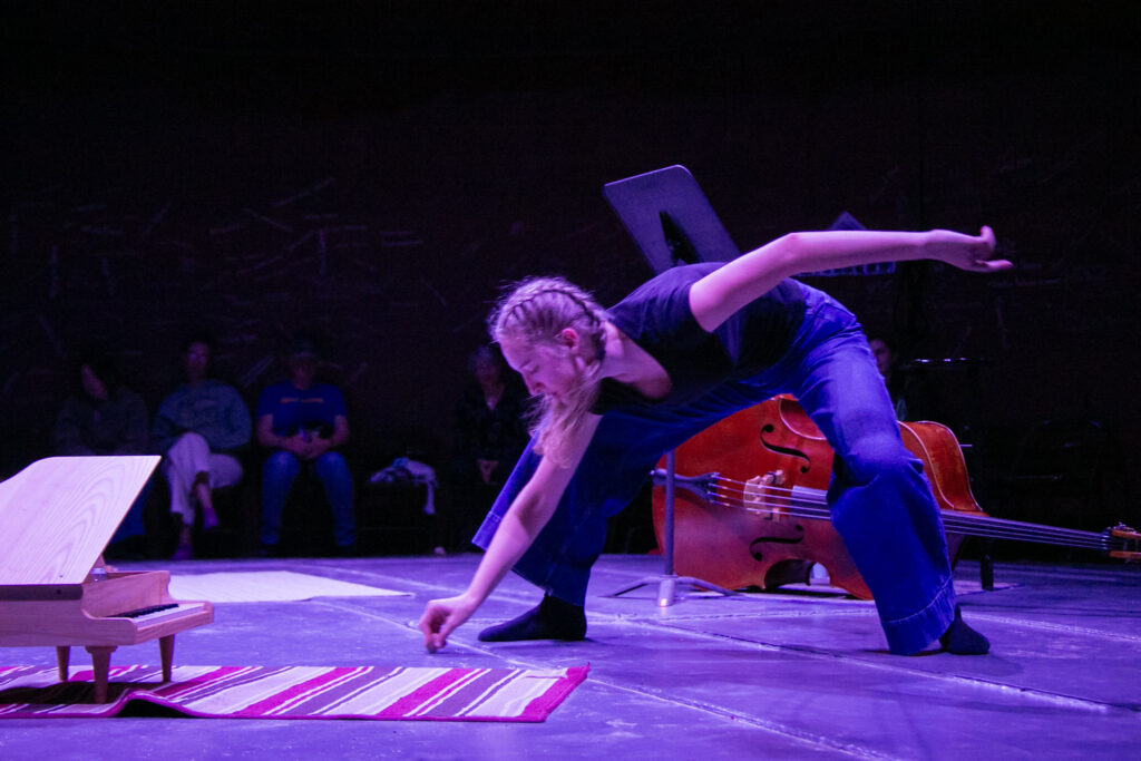 Holly McMahon stands legs wide as she leans down, right hand scraping a rock on the floor, left arm extended behind her. A double bass and toy piano are nearby and an audience shrouded in darkness is barely visible behind her.