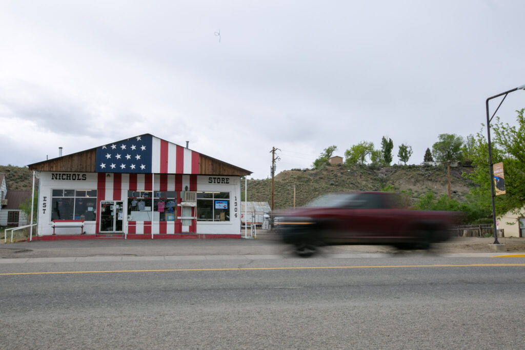 A red pickup truck speeds by a store painted like an American Flag that reads, "NICHOLS STORE."