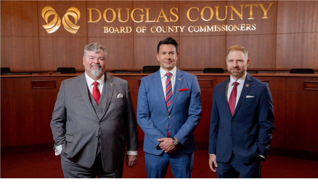 three men in suits standing in Douglas County's commission chambers, stare into the camera