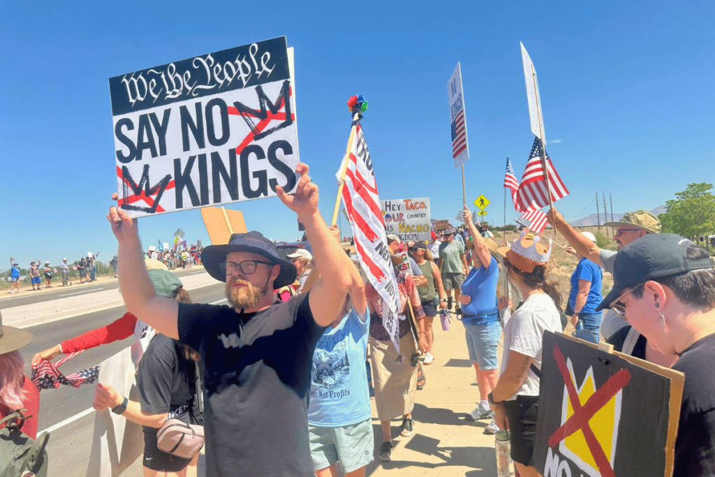 A group of people are gathered on a bridge, holding signs and flags. The signs read "13th Street" and "13th Street No Parking". The flags are red and white, and the people are wearing hats.