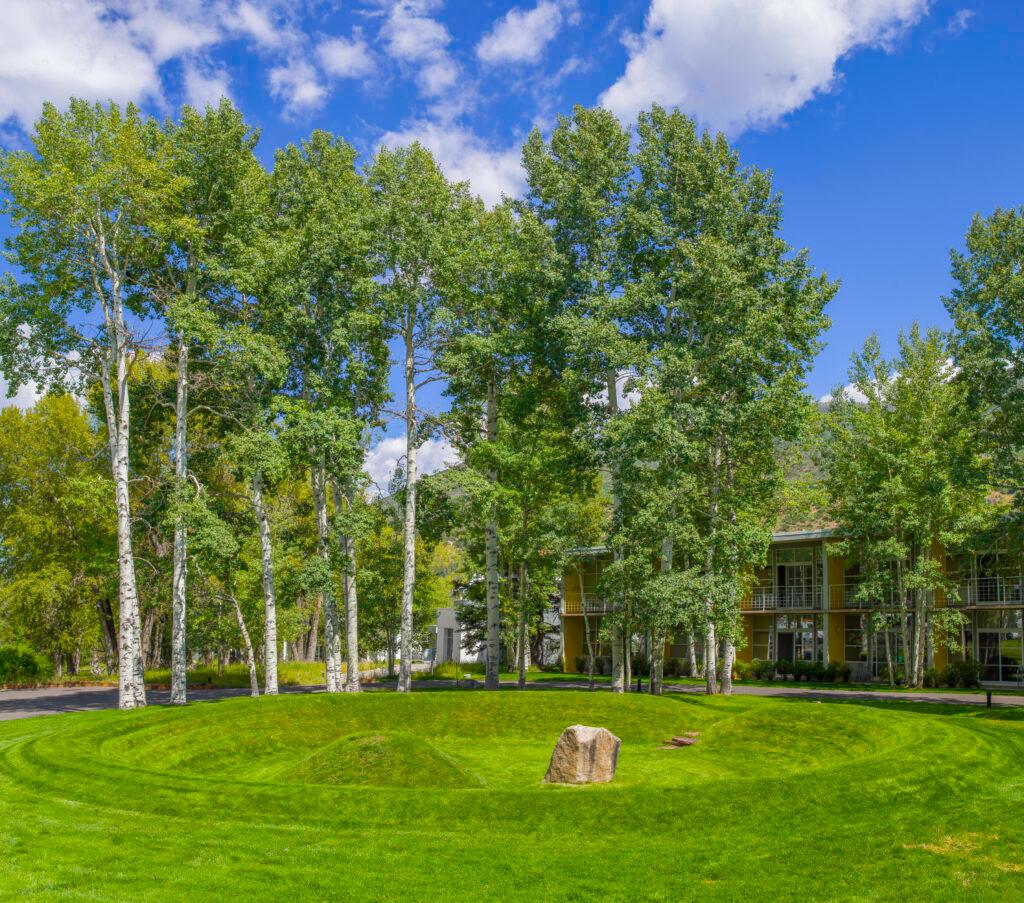 A lawn with a recessed circle. The recessed circle contins a small grass mound and a brown rock. There is one small walking path providing entry into the recessed area.