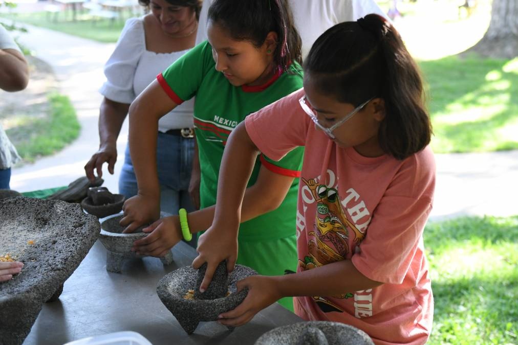 Children learn to grind corn by hand on a metate, a bowl made from stone.