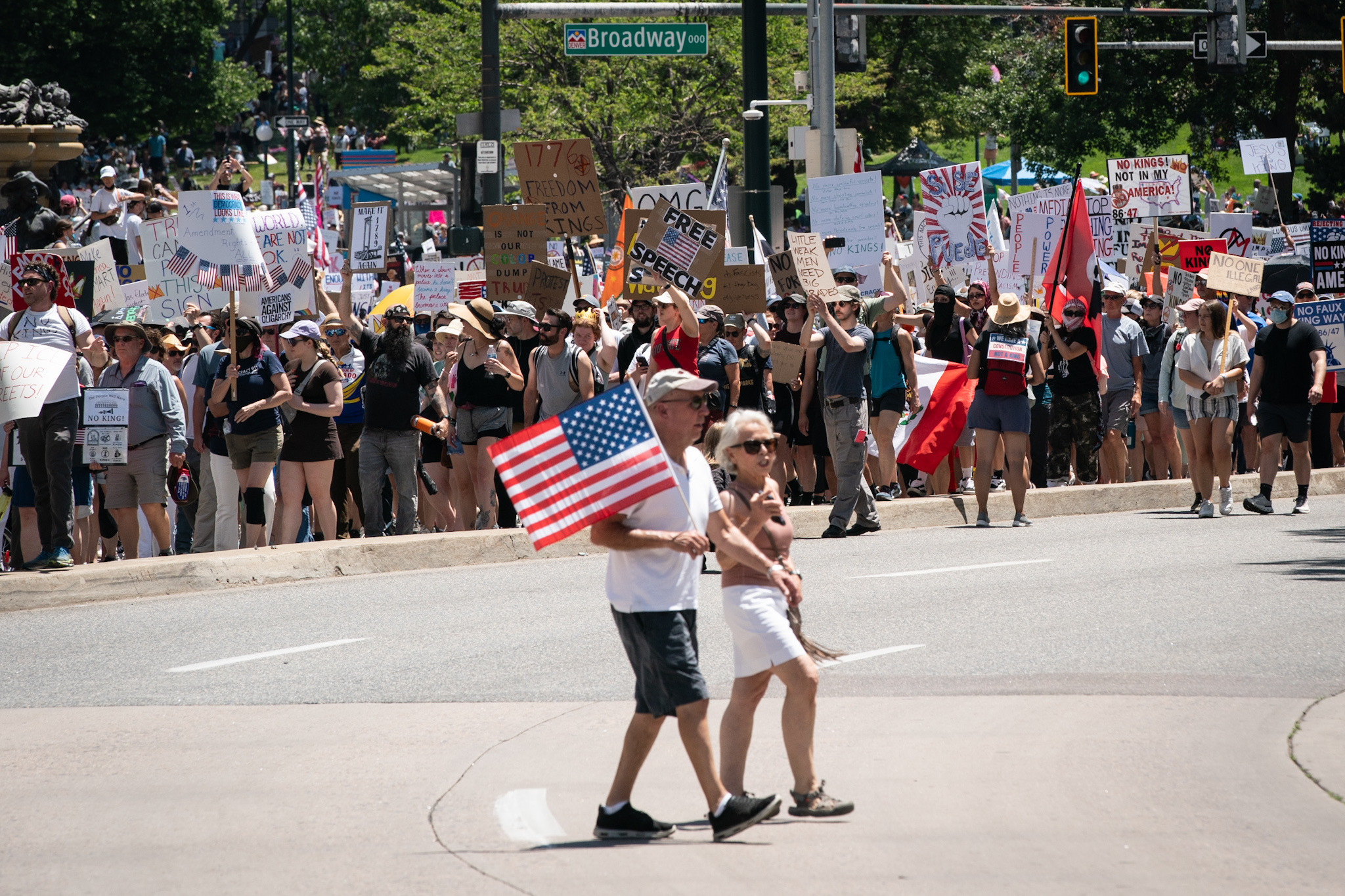 Two people holding an American flag walk across the street while a crowd of protestors stand behind them