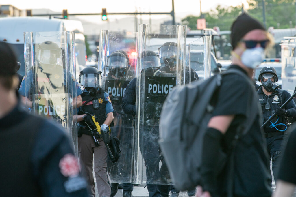Police with riot gear and tall shields walk in background. A protester in sunglasses and a medical mask in the front.