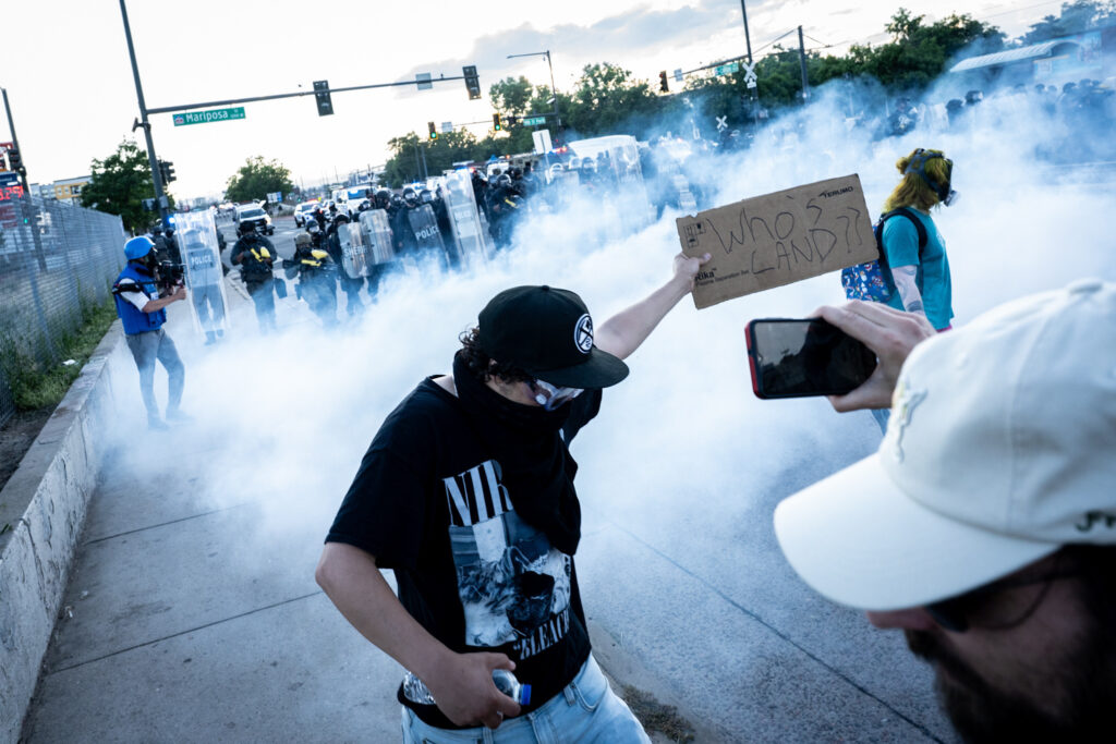 Man in black shirt, mask, hat holds sign that reads "Who's Land?" As smoke surrounds him.