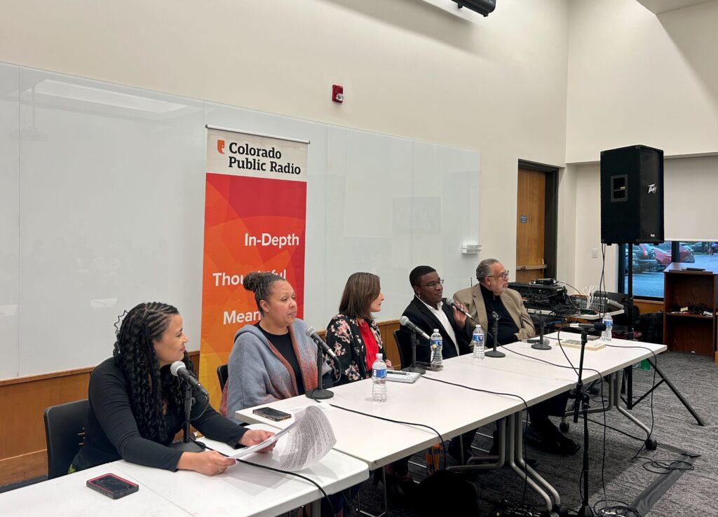 Five people are seated at a table that is diagonal in the photo. There is a vertical banner that is orange behind them that says "Colorado Public Radio" and toward the right is a large audio speaker on a stand.