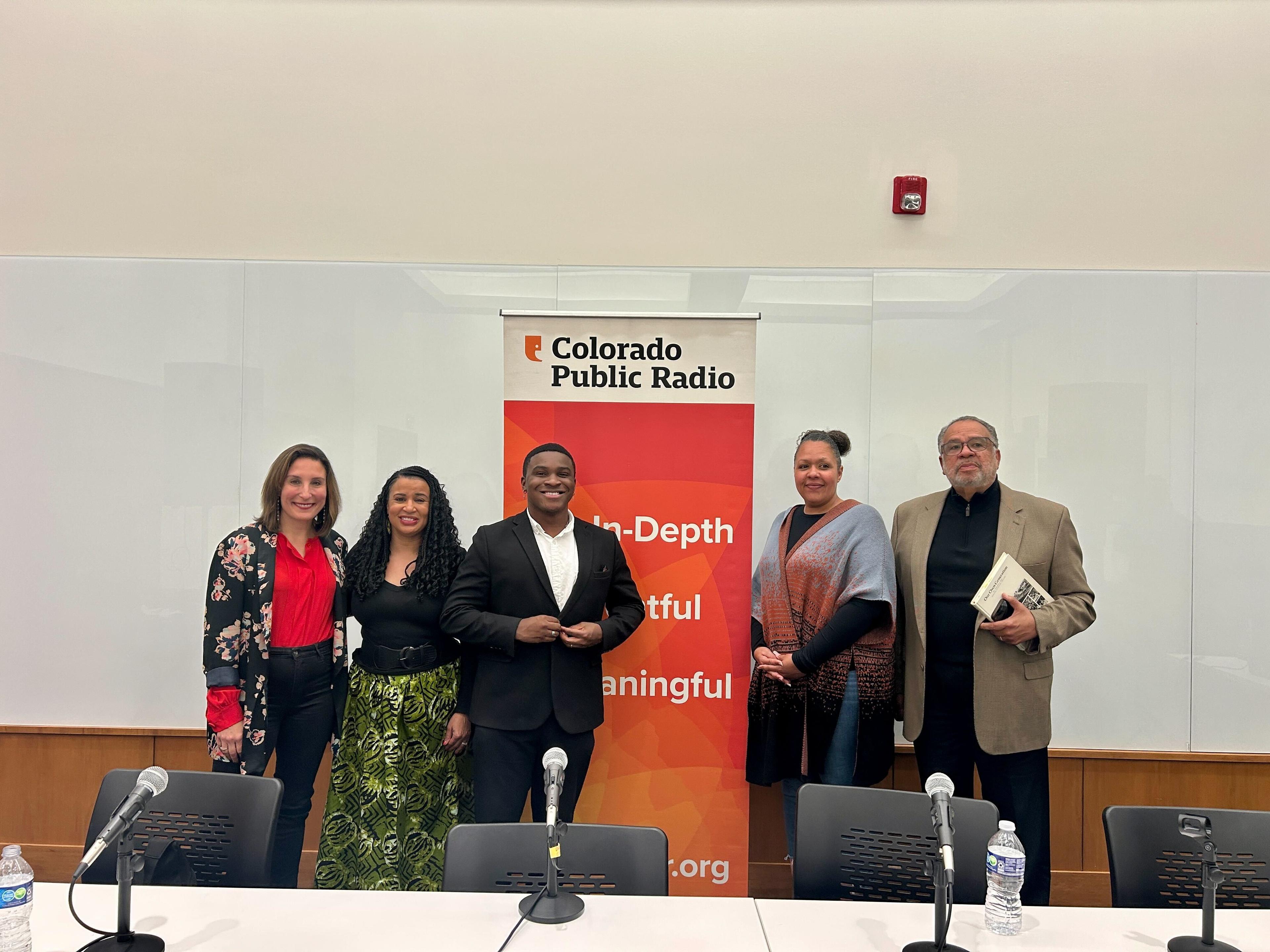 Five people stand in front of an orange banner that says "Colorado Public Radio" at the top. The the foreground is a table with microphones.