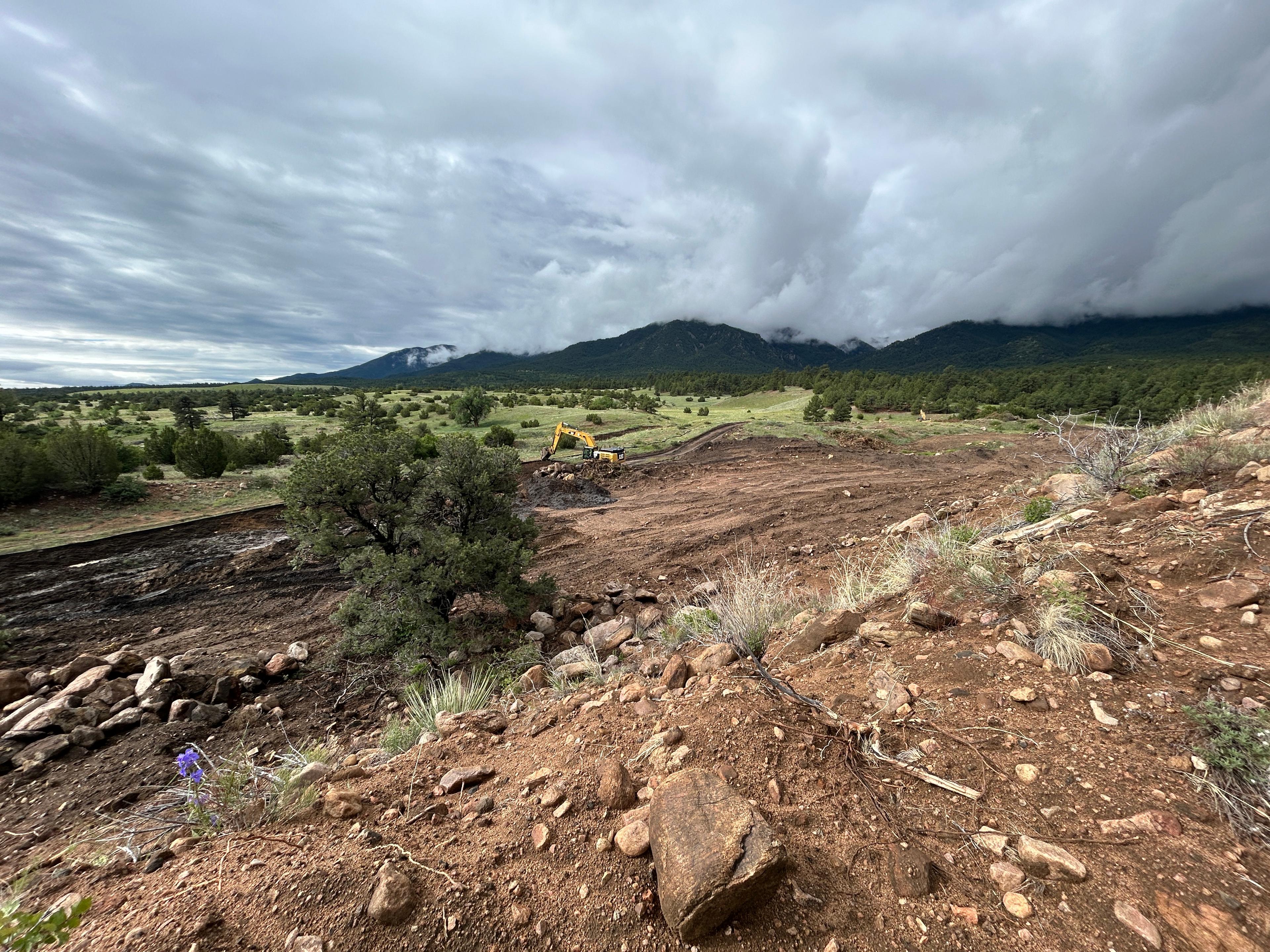 heavy equipment works a large area of disturbed ground with mountains and clouds in the distance