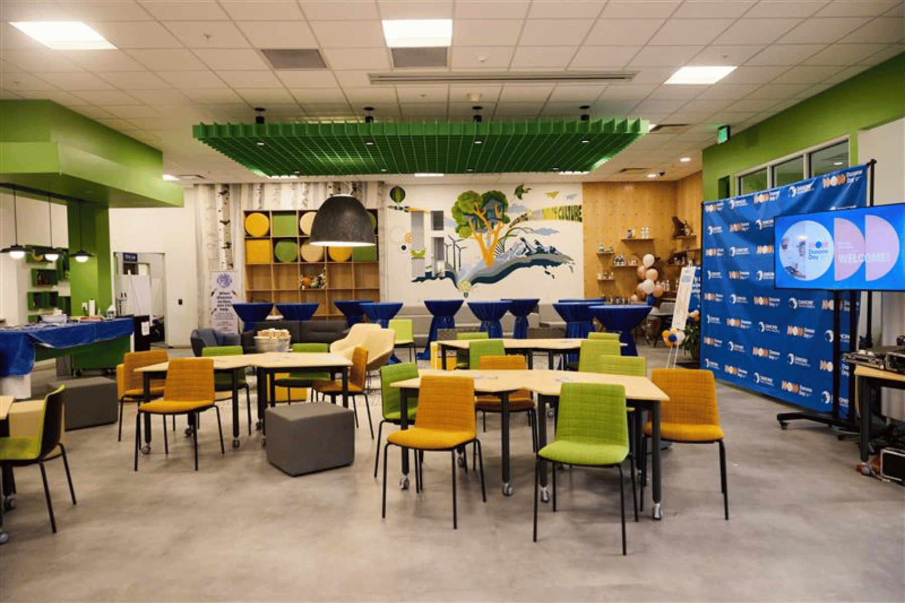 Inside a very modern looking workspace with tables and orange and green chairs, some grey cubes, tables, and stand-up stations