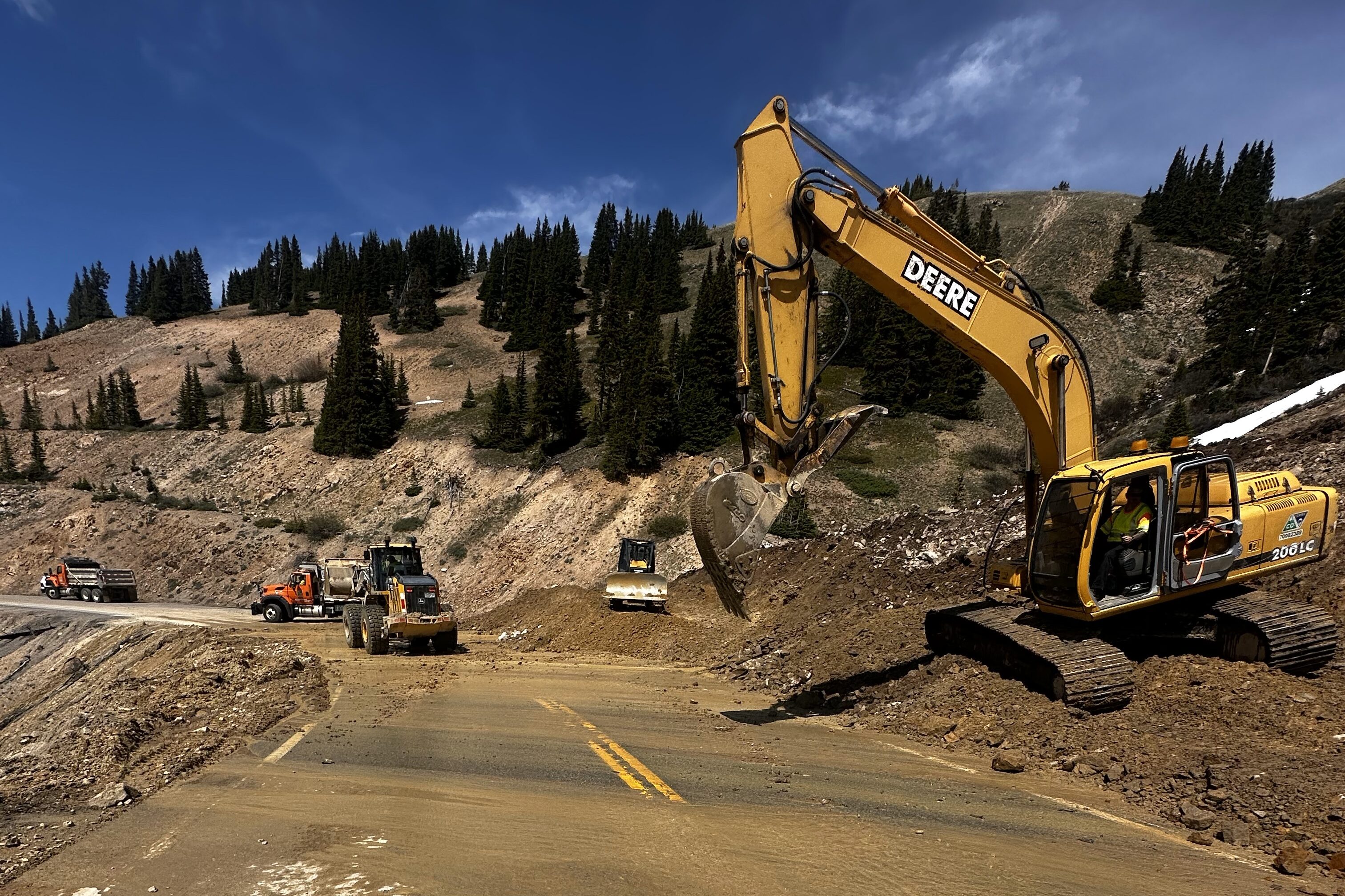 A yellow excavator perched on a pile of mud alongside a two-lane road
