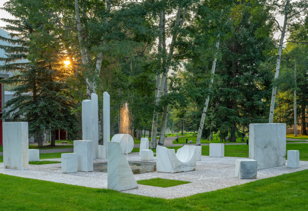 A grass field with a large square inside. The square holds various marble monoliths and other marble shapes.
