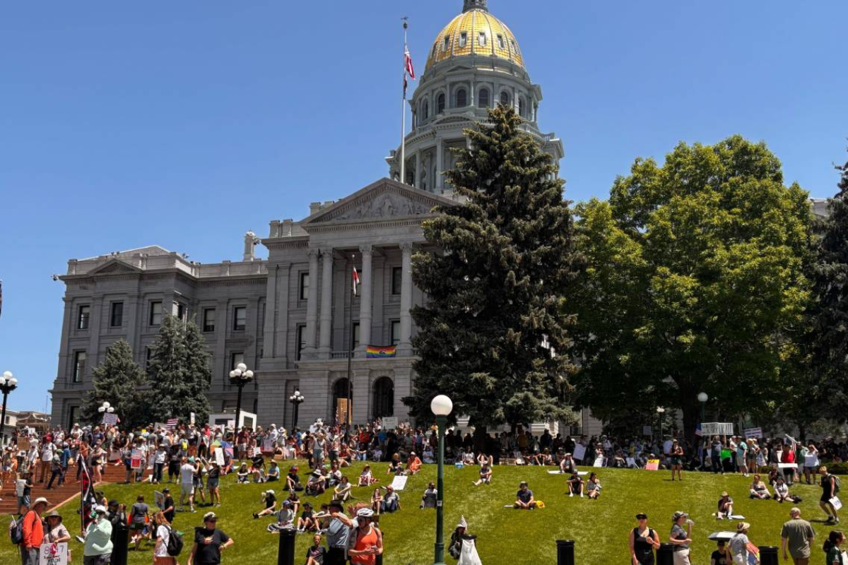 People sit and hold signs up near the Colorado State Capitol with a golden dome.