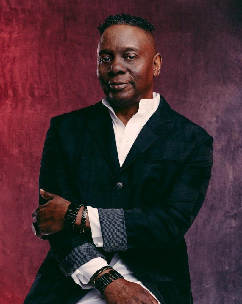 Photo shows a man in a black coat with a white shirt in front of a burgundy background