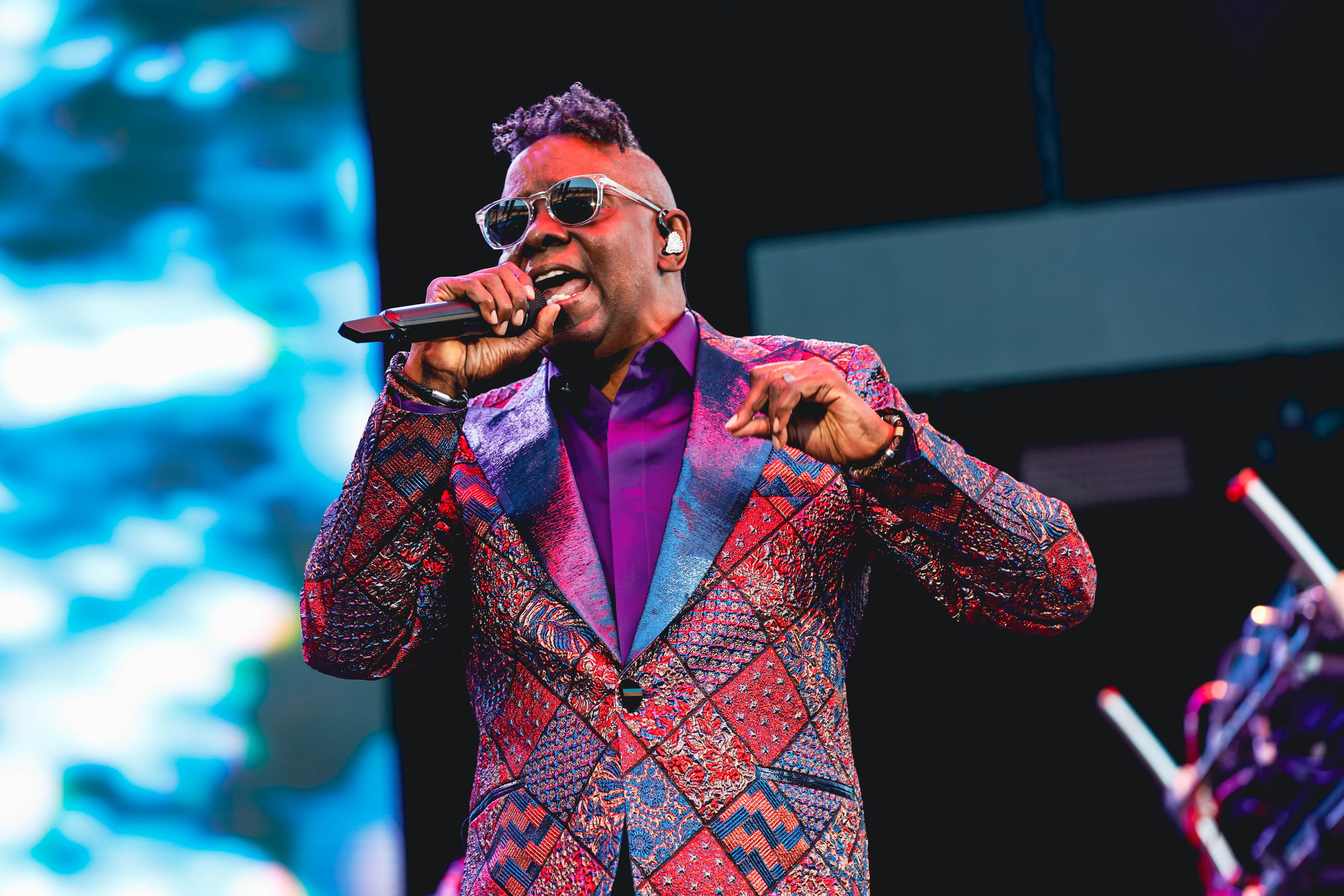 A man with sunglasses in a purple and red plaid suit coat with a purple shirt sings into a microphone on stage