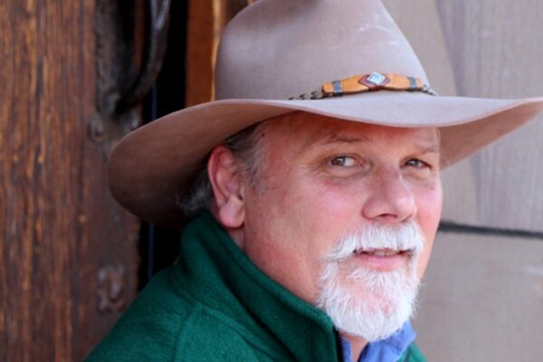 Man is seen in 3/4 profile wearing a cowboy hat and green vest over a blue shirt. He has a white mustache and beard.