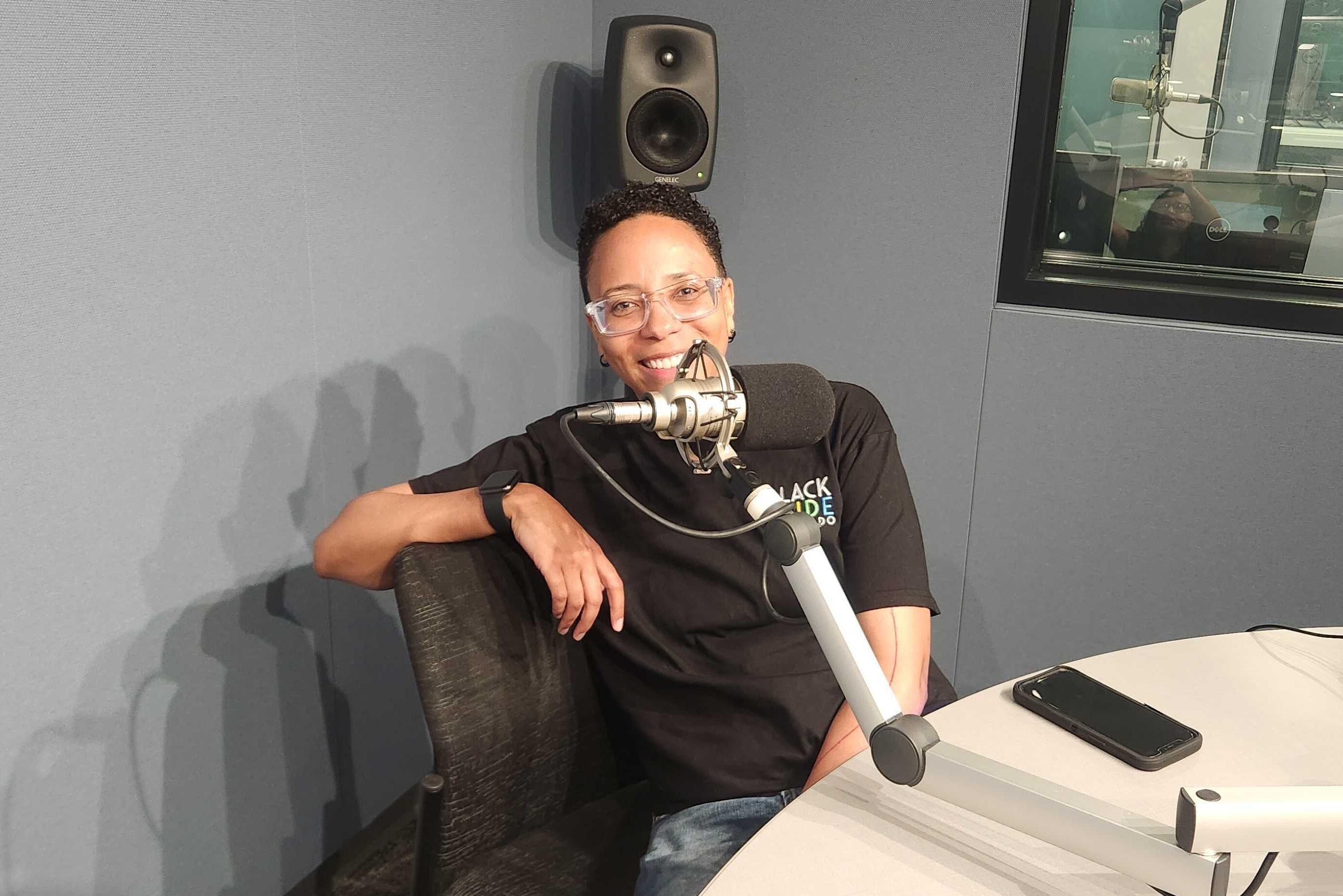 A person with short cropped black hair wearing a black tee shirt sits behind a microphone in the CPR News studio. She is wearing glasses and smiling. Her right arm is resting on the back of the chair.