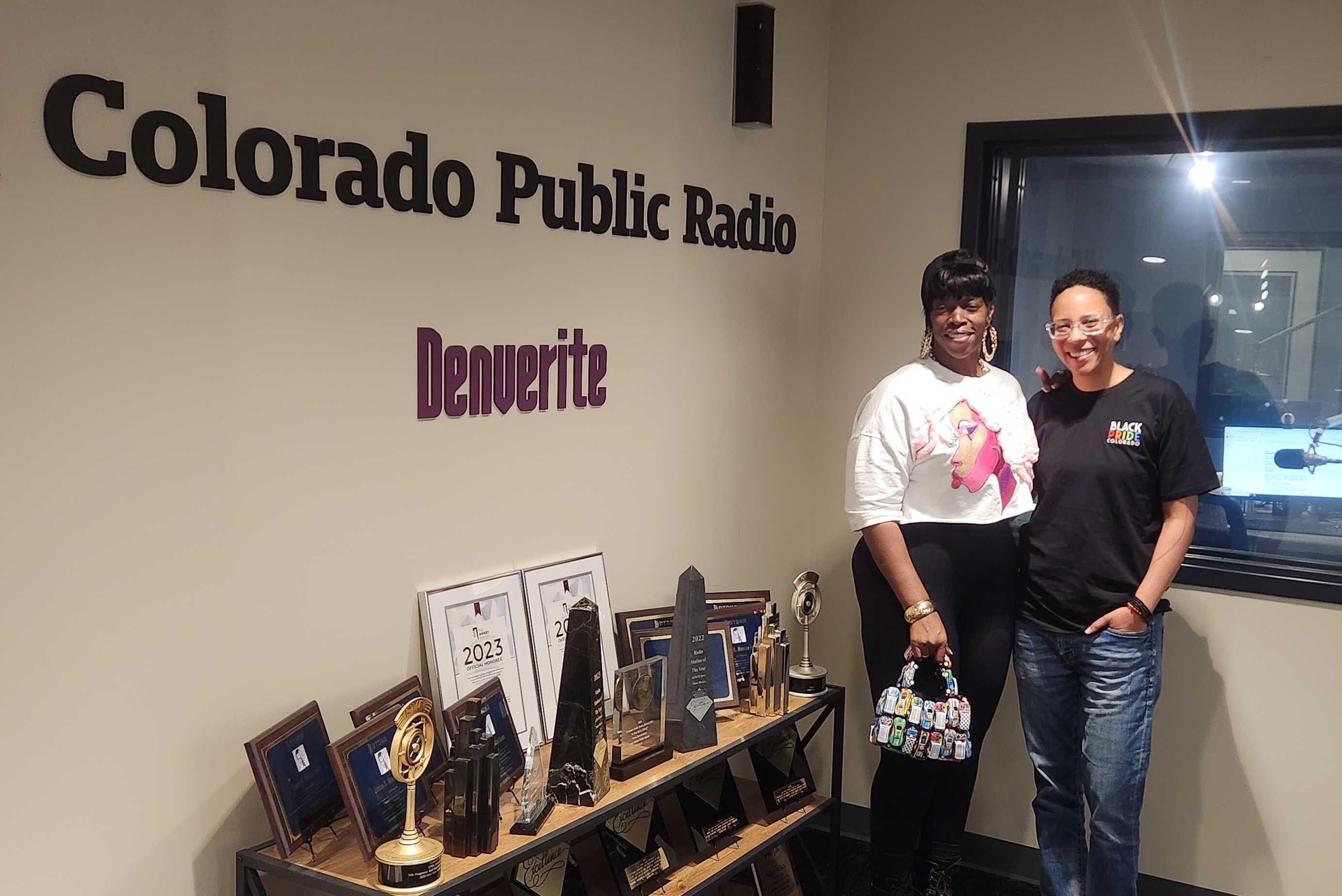 Two people stand in front of a glass studio window next to a sign that says Colorado Public Radio. One is wearing a white shirt with black pants and the other is wearing a black shirt with jeans.