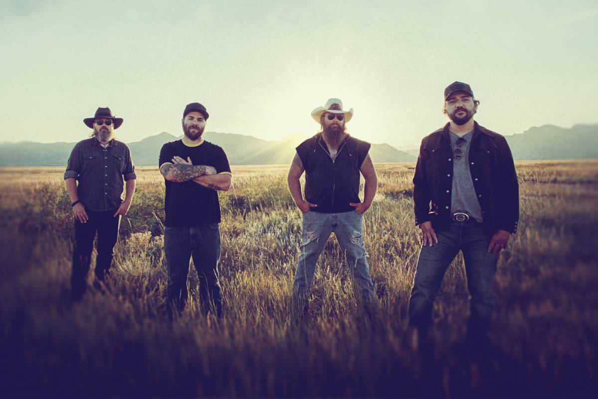 Four men standing in a row in a field of tall grass with mountains in the background. The are all wearing caps or cowboy hats.