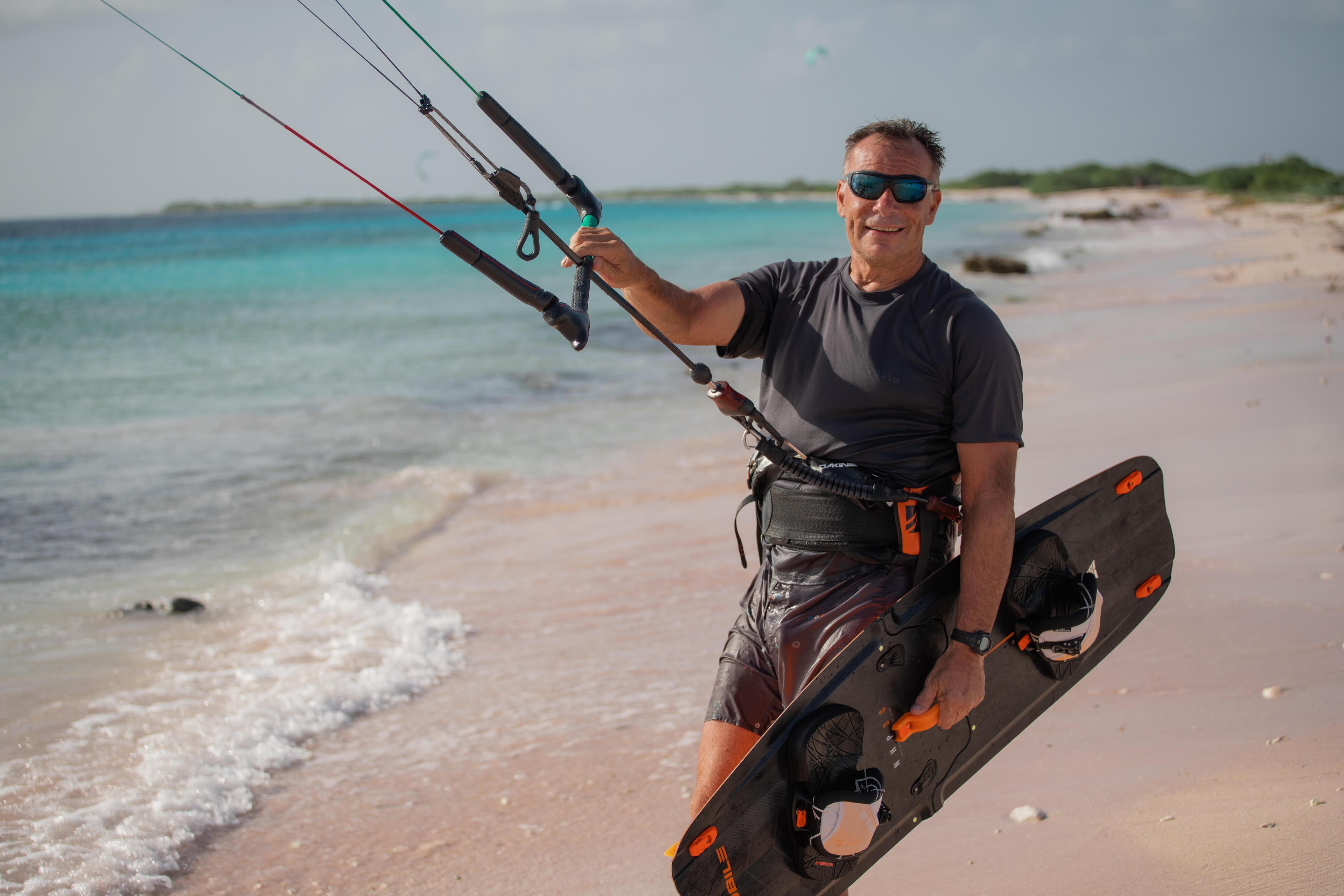 A fit man standing on the beach holding a board and parasail.