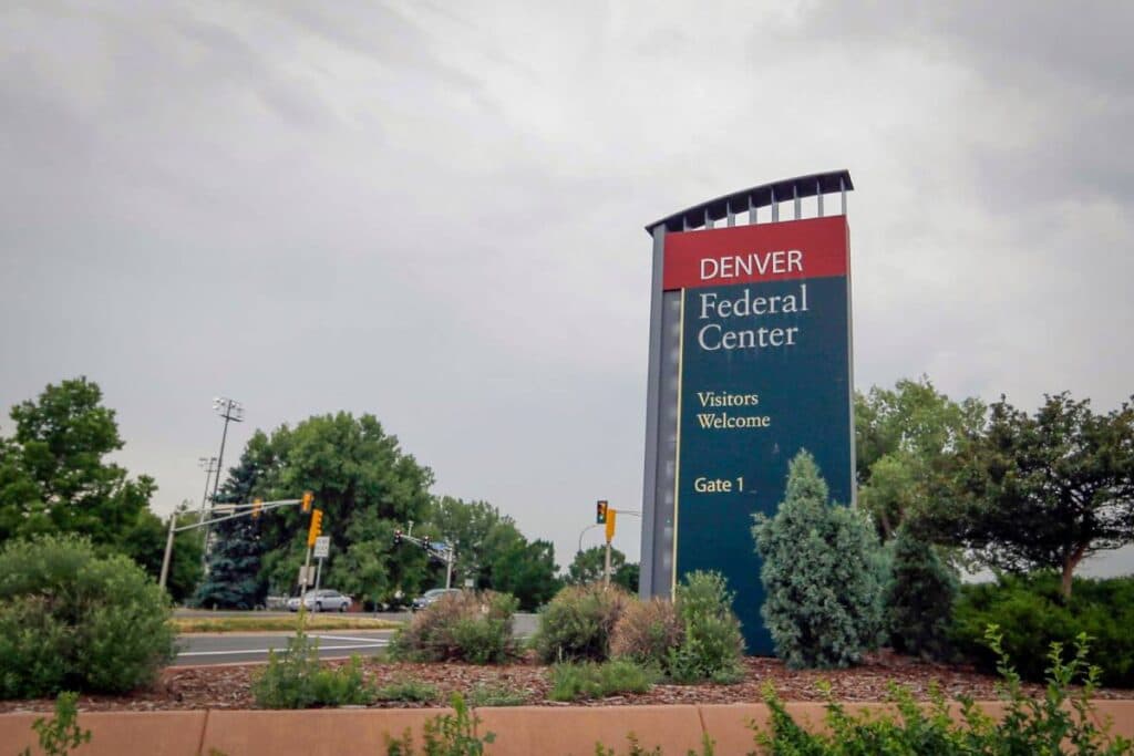 The image features a large sign with the words &quot;Denver Federal Center&quot; written on it. The sign is located in front of a building, and there are trees in the background. The scene is set on a street, and there are cars parked nearby.